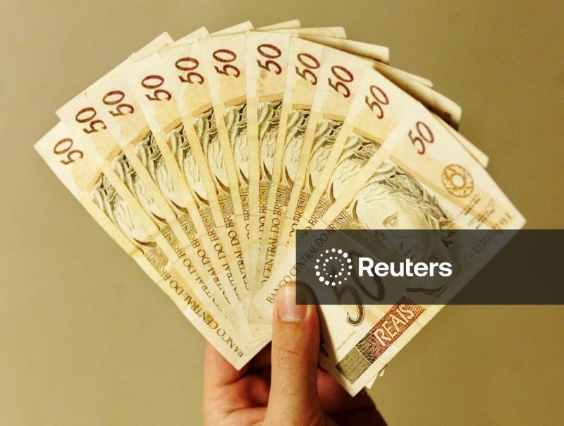 Mão segurando várias cédulas de dinheiro de cinquenta reais do Brasil, formando uma leve curva, em fundo neutro.