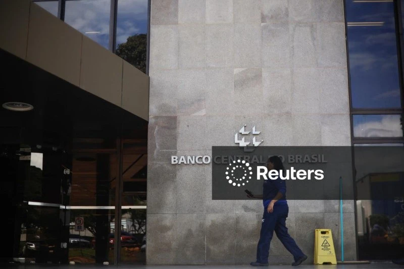 Imagem da fachada do Banco Central do Brasil com fachada moderna, com uma pessoa caminhando na calçada e um aviso de piso molhado na frente do prédio.