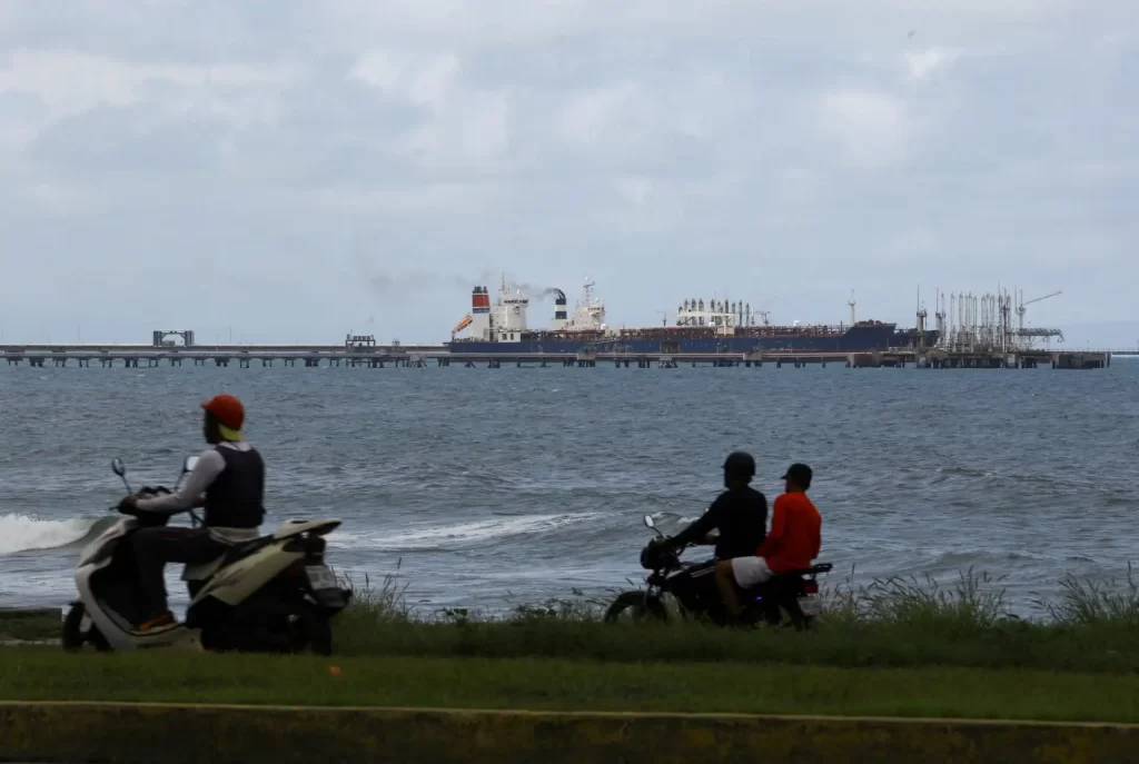 Homem e duas crianças em motos na orla de um litoral com um navio de grande porte ao fundo navegando no mar, céu parcialmente nublado.