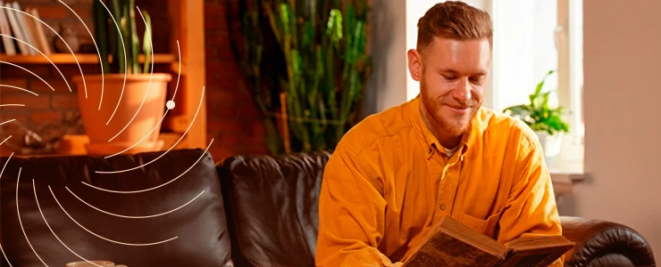Homem lendo um livro sentado em um sofá de couro marrom, ambiente com plantas verdes ao fundo e luz natural entrando pela janela.