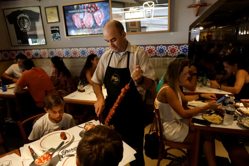Homem preparando churrasco em restaurante movimentado com várias pessoas ao redor, decorado com estilo tradicional brasileiro, durante uma refeição.
