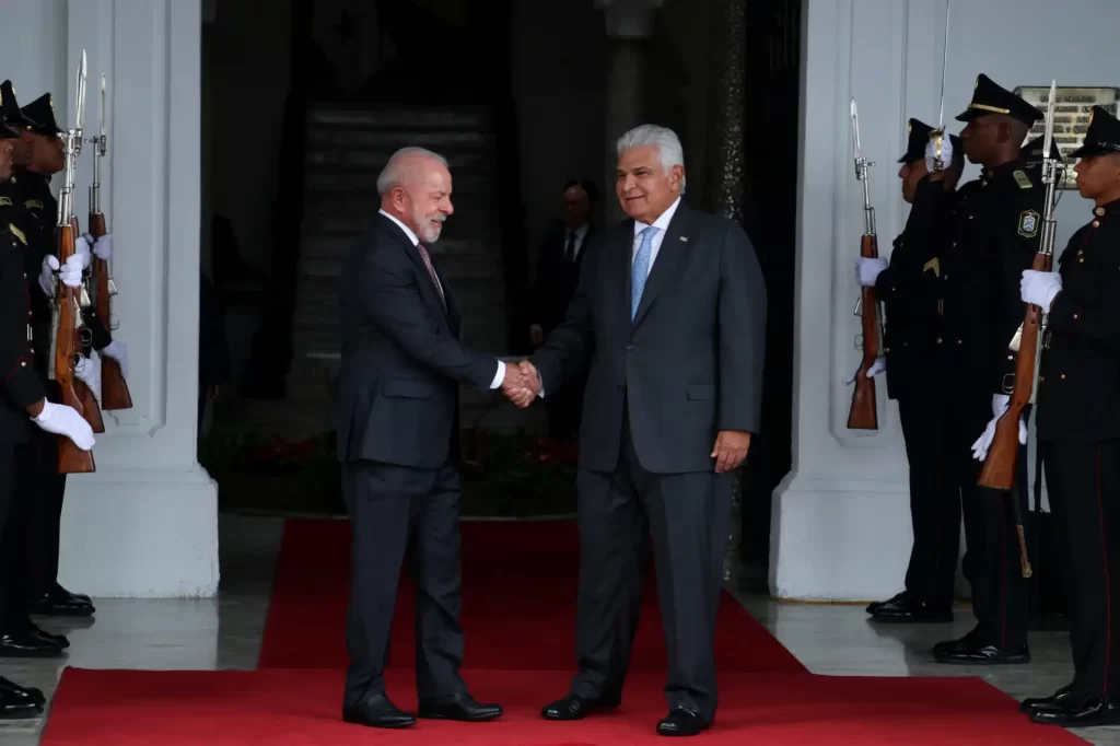 Líderes mundiais se cumprimentando durante uma cerimônia oficial, com policiais uniformizados formando uma guarda de honra ao lado. Momento de diplomacia e cordialidade entre representantes de países.