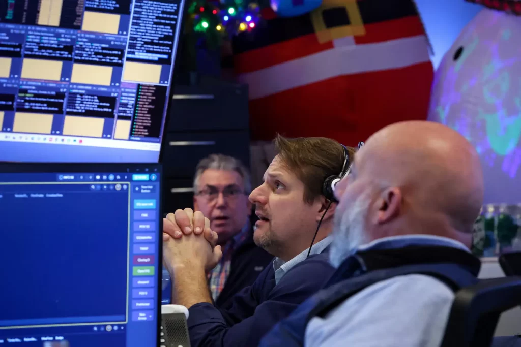 Três operadores de bolsa analisando dados financeiros em uma sala de controle com múltiplos monitores, ambiente de negociação de ações e mercado financeiro ao fundo.