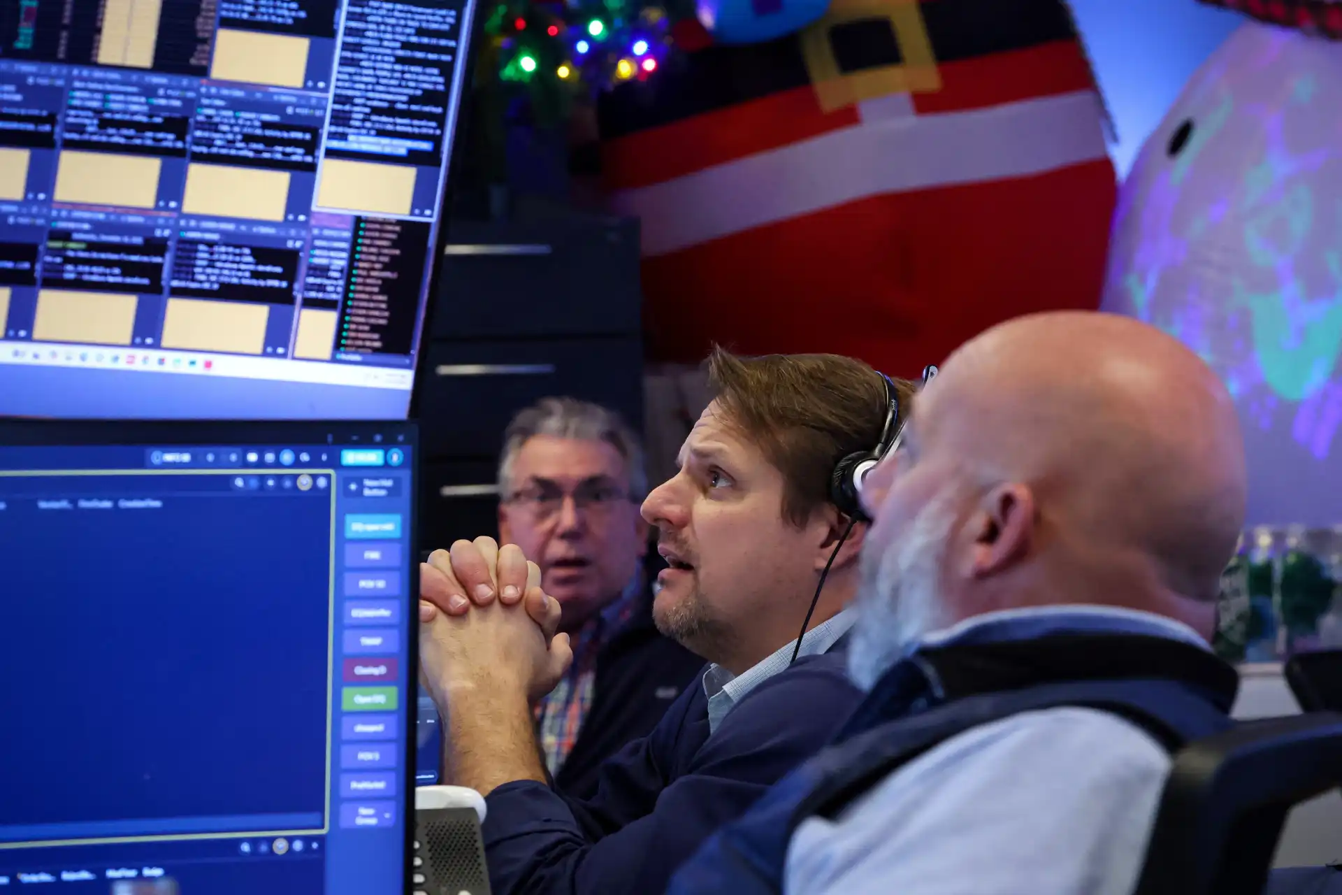 Três operadores de bolsa analisando dados financeiros em uma sala de controle com múltiplos monitores, ambiente de negociação de ações e mercado financeiro ao fundo.