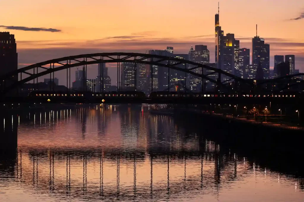 Paisagem urbana de Frankfurt ao entardecer com céu colorido, rio refletindo a cidade e ponte histórica em destaque.
