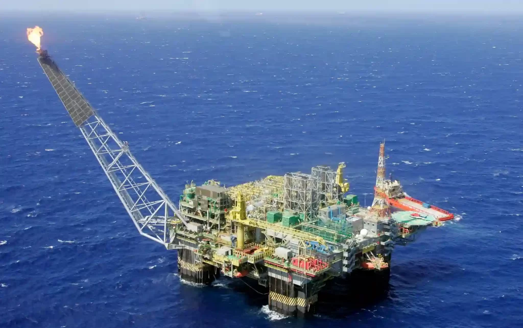 Plataforma de petróleo offshore no oceano, com equipamentos de extração e torre de perfuração, em mar aberto com céu claro ao fundo.
