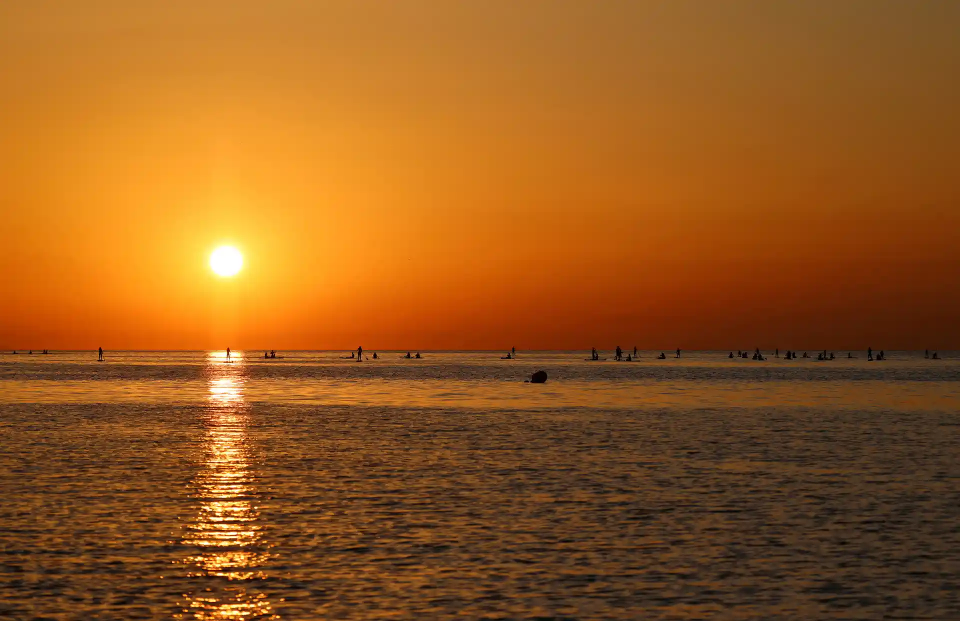 Paisagem de pôr do sol na praia com céu laranja e reflexo no mar, pessoas e barcos ao fundo, criando uma cena tranquila e bonita.