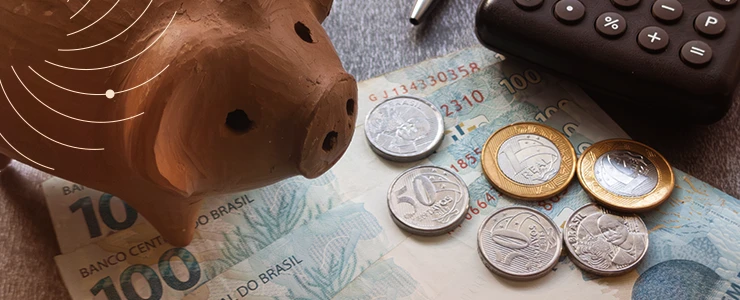 Porquinho de cerâmica, moedas de real e nota de dinheiro brasileiro com calculadora ao fundo, representando economia e finanças pessoais no Brasil.
