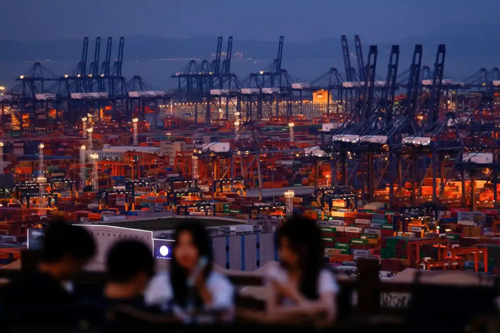Imagem de um porto de containers à noite, com várias torres de crane e iluminação, vista ao fundo com três pessoas ao telefone em primeiro plano.