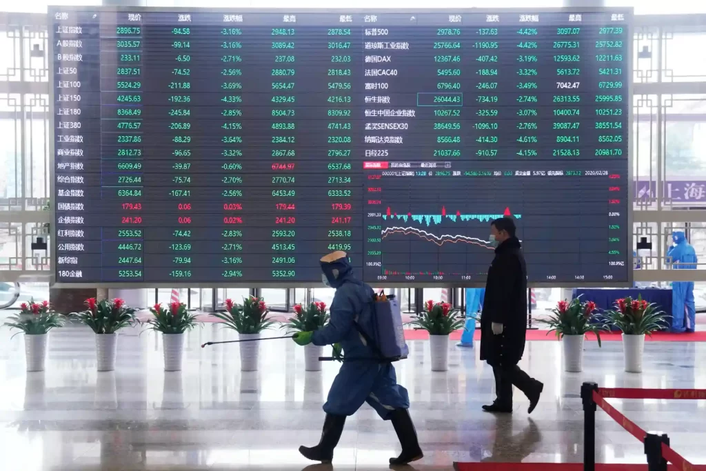 Imagem de uma tela gigante de indicadores financeiros com várias ações e índices de mercado na bolsa de valores, enquanto duas pessoas caminham no local. A cena reflete o movimento do mercado financeiro na China.