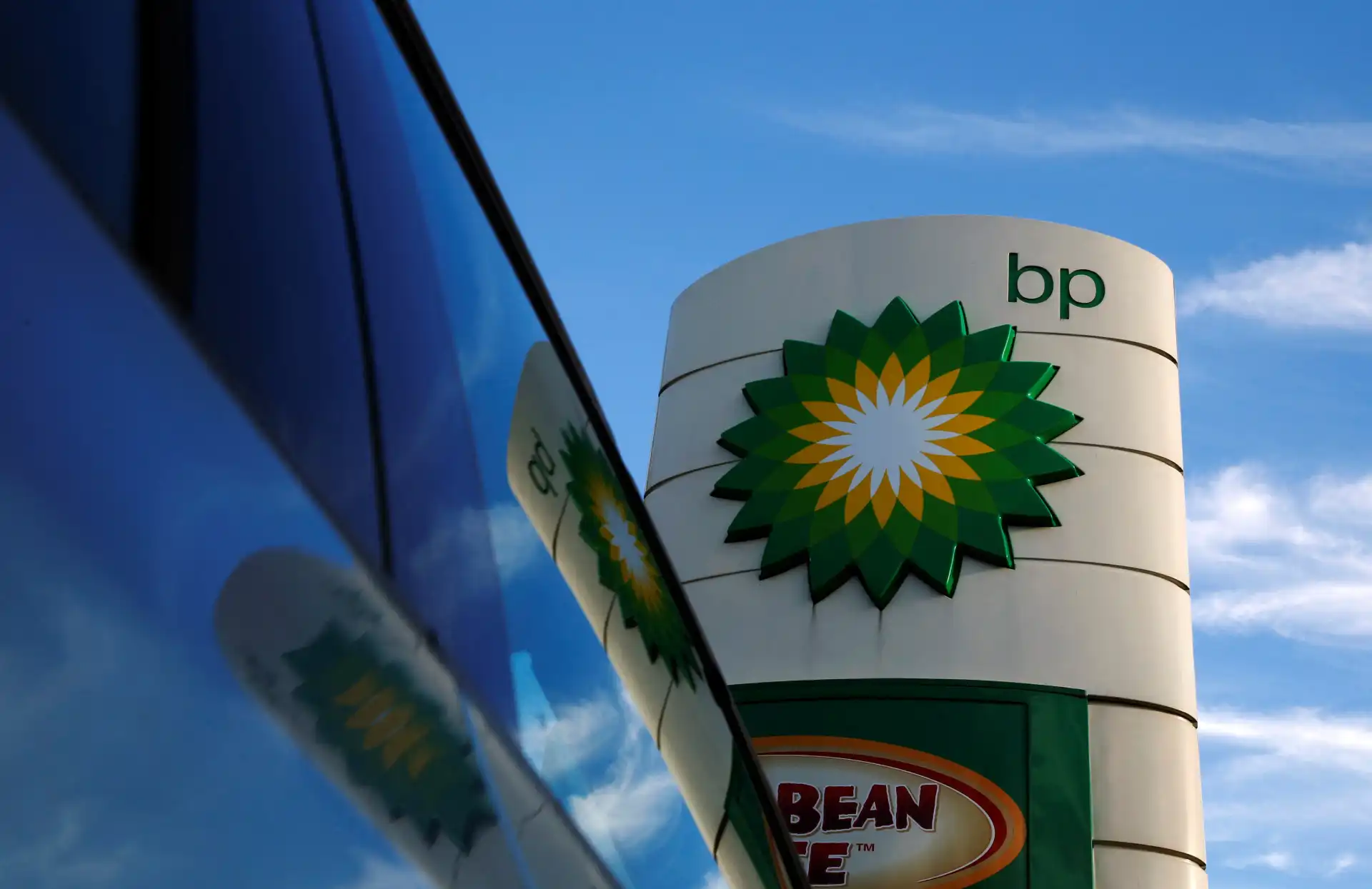 Torre de bombas de combustível da BP refletida na janela de um carro, com céu azul ao fundo, destaque para o logo da BP.