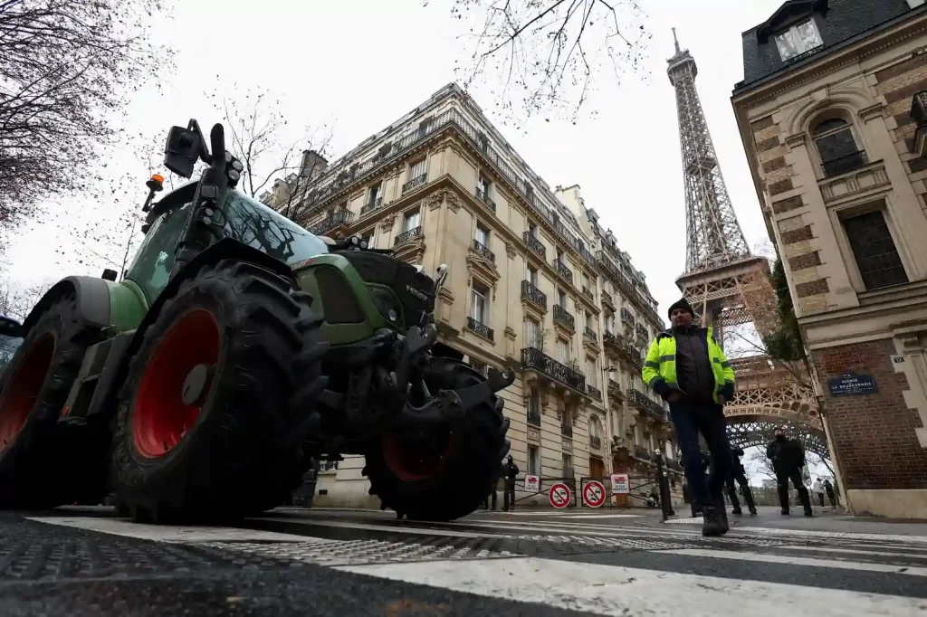 Imagem de uma rua em Paris com o Eiffell Tower ao fundo, um trator agrícola de cor verde e detalhes vermelhos na frente e um policial de colete refletivo ao lado, comemorando a paisagem urbana com edifícios clássicos e árvores sem folhas.