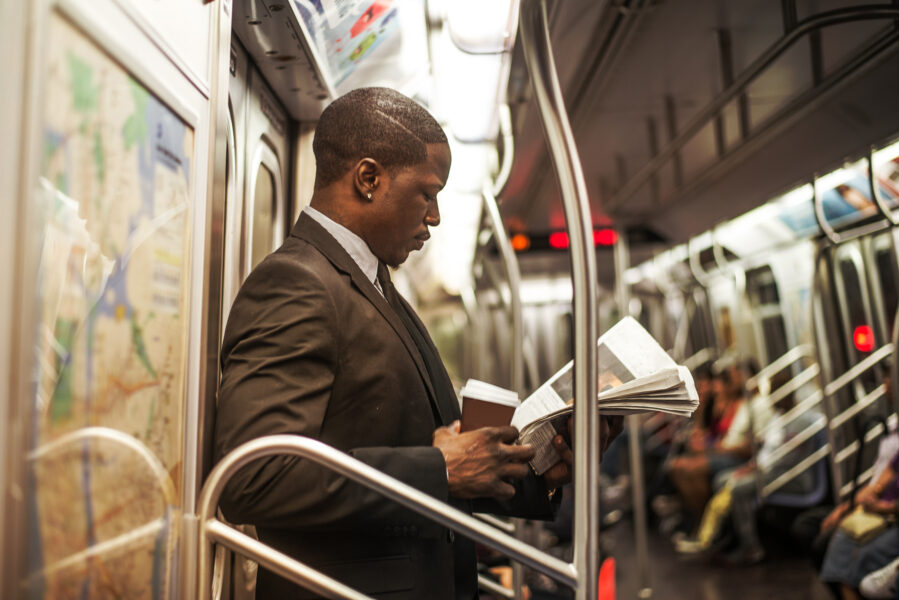 Uma pessoa em pé dentro de um vagão de metrô lê um jornal enquanto segura um copo de café.