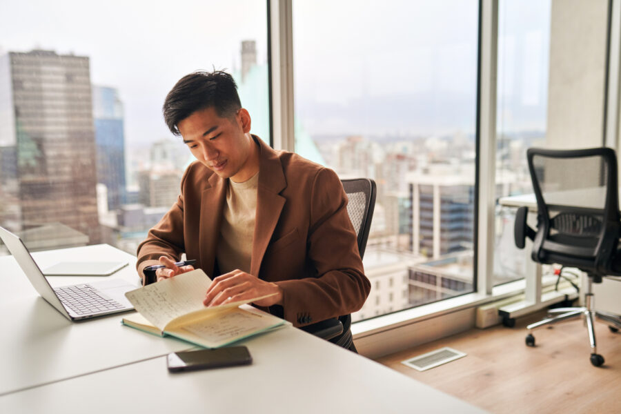 Pessoa sentada a uma mesa com um livro e um laptop em um escritório em um prédio alto, com vista para a cidade.